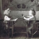 2 małe dziewczynki siedzą przy niskim stoliku, naprzeciwko siebie. Każda z nich czyta książeczkę.