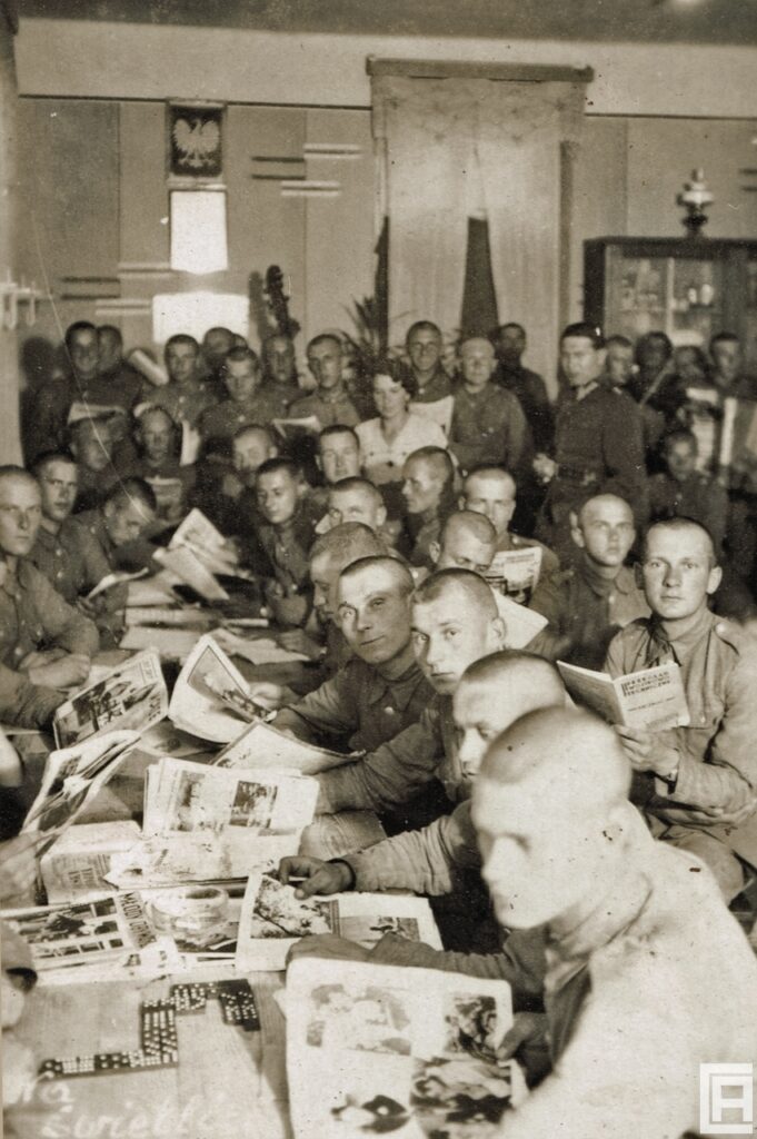 W dużej sali wokół stołu siedzą ciasno obok siebie żołnierze. Każdy z nich przegląda, czyta czasopismo lub książkę.