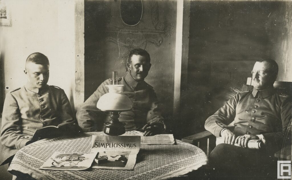 Trzech żołnierzy w mundurach z pierwszej wojny światowej siedzi przy stole, na którym stoi ozdobna lampa naftowa i leży czasopismo "Simplicissimus" z 1 czerwca 1915 roku.