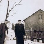 Na fotografii jest bałwan, po dwóch jego stronach stoją mężczyźni. W tle widać drewniany płot i budynek mieszkalny. Fotografia zrobiona zimą.