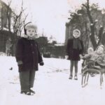 Na ulicy pełnej śniegu dwoje dzieci (chłopiec i dziewczynka) stoją trzymając sznurki od sanek, dwoje małych dzieci siedzi na sankach. W tle widać budynki.