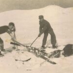 Trzech mężczyzn na nartach. Jeden z nich się wywrócił do śniegu, pozostali starają się mu pomóc wstać. Wokół dużo śniegu.