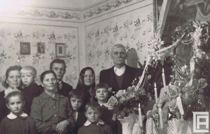 Zdjęcie rodziny wielopokoleniowej, przedstawia 10 osób. Z tyłu widoczne zdjęcie komunijne. Z prawej widoczna rama świętego obrazu.