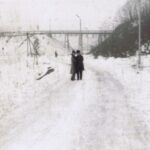 Dwie osoby pozują do zdjęcia na ścieżce. Teren wokół jest pusty i zaśnieżony, daleko z tyłu widać postać człowieka, rury, słupy.