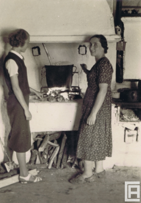 Dwie młode kobiety stoją po dwóch stronach kuchennego paleniska. Na rozpalonych drwach stoi sporych rozmiarów kociołek z chochlą.