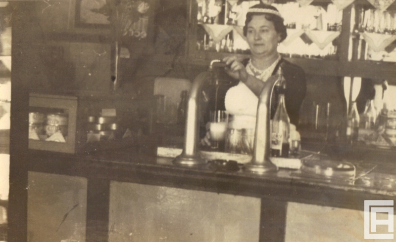 Za ladą barową stoi kobieta nalewająca z dystrybutora piwo do kufla. W tle widoczne szklanki, butelki.