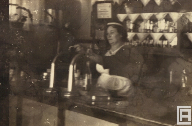 Za ladą barową stoi kobieta nalewająca z dystrybutora piwo do kufla. W tle widoczne szklanki, butelki, kufle.