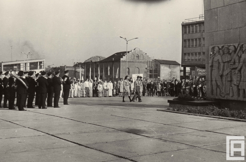 Wokół placu zebrana jest grupa ludzi: orkiestra zakładowa, osoby w kostiumach. Dwoje harcerzy idzie w kierunku pomnika z kwiatami. W tle widoczny jest budynek synagogi.