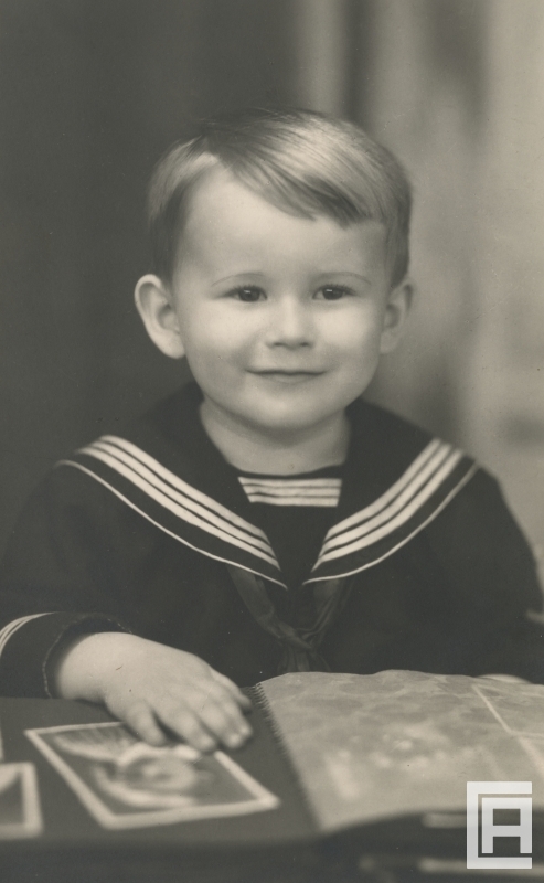 Uśmiechnięty chłopiec w ubranku marynarskim trzyma prawą dłoń na otwartym albumie ze zdjęciami.