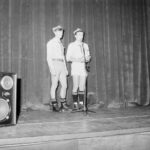Dwóch harcerzy stoi na scenie przy mikrofonie. Obok duży głośnik.
