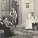 Fotografia przedstawia scenę, salonik. Kobieta z mężczyzną siedzą na kanapie, naprzeciw inna para przy krześle, do pomieszczenia wszedł trzeci mężczyzna. Stroje XIX-wieczne.