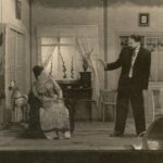 Fotografia przedstawia scenę, salonik. Kobieta siedzi na fotelu, obok mężczyzna gestykuluje, rozmawiają.