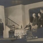 Fotografia przedstawia scenę. Trzy kobiety i dwóch mężczyzn w pokoju z niemieckim żołnierzem.