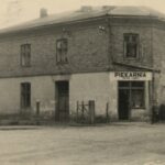 Fotografia przedstawia jednopiętrowy budynek z wejściem narożnym do piekarni. U góry pięć okien, na dole trzy. Część górna budynku ceglana.