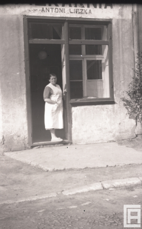 Fotografia przedstawia kobietę w białym fartuchu stojącą przed drzwiami.