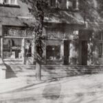 Fotografia przedstawia witrynę biblioteki wraz z tabliczką, obok drzwi. Widoczne otoczenie, chodnik i drzewa.