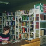 Fotografia przedstawia bibliotekarkę przeglądającą gazetę, za nią widoczne regały z książkami.