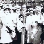 Fotografia przedstawia grupę osób otaczającą chłopca trzymającego flagę na drzewcu. Kobiety ubrane są w stroje służbowe pielęgniarek.