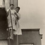 Fotografia przedstawia dwie kobiety w białych fartuchach stojące na stopniu metalowych schodów.