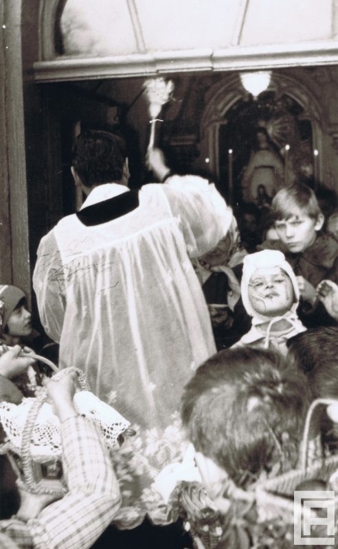 Fotografia przedstawia księdza z kropielnicą oraz tłum dzieci z koszyczkami wielkanocnymi.