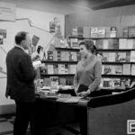 Fotografia przedstawia kobietę przy stoisku z książkami i prasą, która rozmawia z mężczyzną.