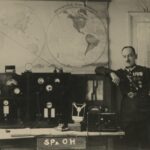 Fotografia przedstawia mężczyznę w mundurze opartego o urządzenie radiowe. W tle wizerunki półkuli północnej i południowej.