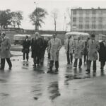 Fotografia przedstawia grupę osób w garniturach i płaszczach przechodzących przez plac w zajezdni autobusowej. W tle widoczny budynek oraz autobusy.