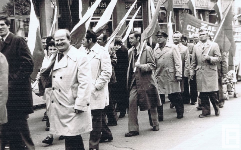 Fotografia przedstawia grupę ludzi z flagami narodowymi, którzy idą w pochodzie. W tle widoczne szyldy sklepowe.