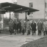 Fotografia przedstawia grupę osób w garniturach i płaszczach przechodzących obok wiaty autobusowej. W tle widoczny budynek.