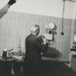 Fotografia przedstawia mężczyznę z kamerą w pomieszczeniu technicznym. Za nim stoi dźwiękowiec z mikrofonem, z prawej wypowiada się pracownik techniczny.