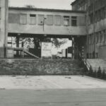 Fotografia przedstawia budynek z bramą i szlabanem. Widoczna ciężarówka i tablica z napisem "Do widzenia".