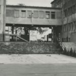 Fotografia przedstawia budynek z bramą i szlabanem. Widoczna ciężarówka i tablica z napisem "Do widzenia".