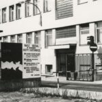 Fotografia przedstawia budynek z bramą wjazdową i szlabanem. Widoczna tablica "Nasz udział w budowie Huty Katowice".