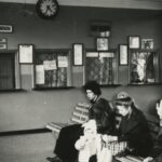 Fotografia przedstawia cztery osoby siedzące w poczekalni autobusowej na ławce: mężczyznę, matkę z dzieckiem i starszą kobietę. Na ścianie okienka do zakupu biletów, zegar oraz różnego rodzaju tablice informacyjne, w tym z napisami „Palenie wzbronione” oraz „ Prosimy o zachowanie czystości”.