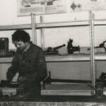 Fotografia przedstawia dwóch mężczyzn w warsztacie. Z tyłu półki z narzędziami.