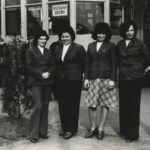 Fotografia przedstawia na pierwszym planie cztery kobiety. W tle widoczni inni ludzie stojący przed budynkiem. Na szybie napis „Dyżurny ruchu”.
