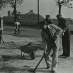 Fotografia przedstawia grupę osób przy taczkach z łopatami. Z tyłu widoczny garbus.