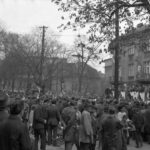 Fotografia przedstawia tłum ludzi podczas pochodu pierwszomajowego. Na budynku widoczny plakat z Włodzimierzem Leninem.