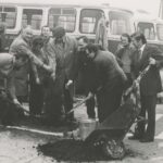 fotografia przedstawia grupę osób z taczkami i łopatami. Mężczyzna w kaszkiecie pochylony jest nad właśnie sadzonym drzewkiem. Z tyłu widoczny autobus.