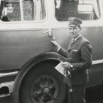 Fotografia przedstawia kierowcę w mundurze otwierającego drzwi autobusu. W wolnej dłoni trzyma proporczyk i kwiat.