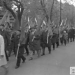 Fotografia przedstawia uczestników pochodu pierwszomajowego, głównie mężczyzn. Z grupy wyróżnia się dziecko w czapce.