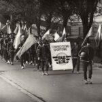 Fotografia przedstawia pochód grupy młodzieży w strojach sportowych, niosą flagi narodowe. Z przodu tablica „Towarzystwo Sportowe Fablok”.