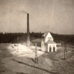 Fotografia przedstawia teren zakładu wydobywczego. Widoczny komin i dwa białe budynki, z tyłu ściana lasu.