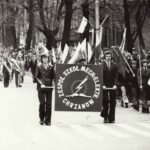 Fotografia przedstawia pochód pierwszomajowy. Na pierwszym planie dwóch mężczyzn niesie tablicę „Zespół Szkół Mechanicznych Chrzanów”.