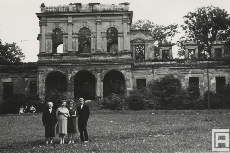 Fotografia przedstawia cztery osoby na pierwszym planie i trzy na dalszym. W tle widoczne ze szczegółami ruiny barokowo-klasycystycznego pałacu.