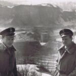 Fotografia przedstawia na pierwszym planie dwóch mężczyzn w mundurach. W tle widoczny teren zakładu wydobywczego.