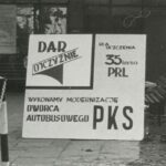 Fotografia przedstawia tablicę z napisami: Dar ojczyźnie. Dla uczczenia 35-lecia PRL. Wykonamy modernizację dworca autobusowego PKS.