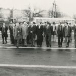 Fotografia przedstawia grupę mężczyzn w garniturach i płaszczach, którzy idą przez zajezdnię autobusową. Z tyłu widoczne bloki i autobusy.