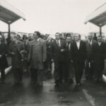 Fotografia przedstawia grupę mężczyzn w garniturach i płaszczach, którzy idą przez zajezdnię autobusową.