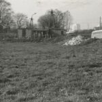 Fotografia przedstawia widok na trawiastą okolicę z kilkoma zniszczonymi budynkami. Z boku widoczne autobusy.